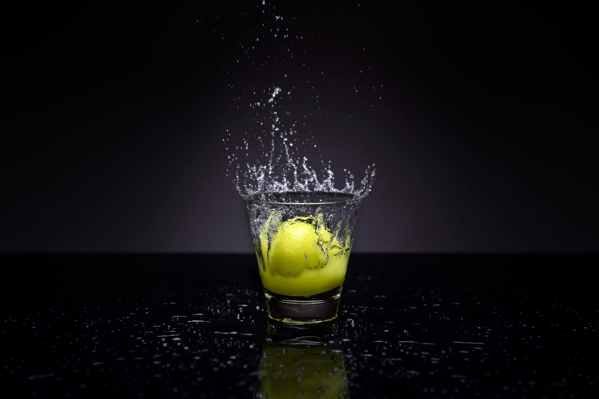 A lemon making a splash in a glass of water against a dark background.